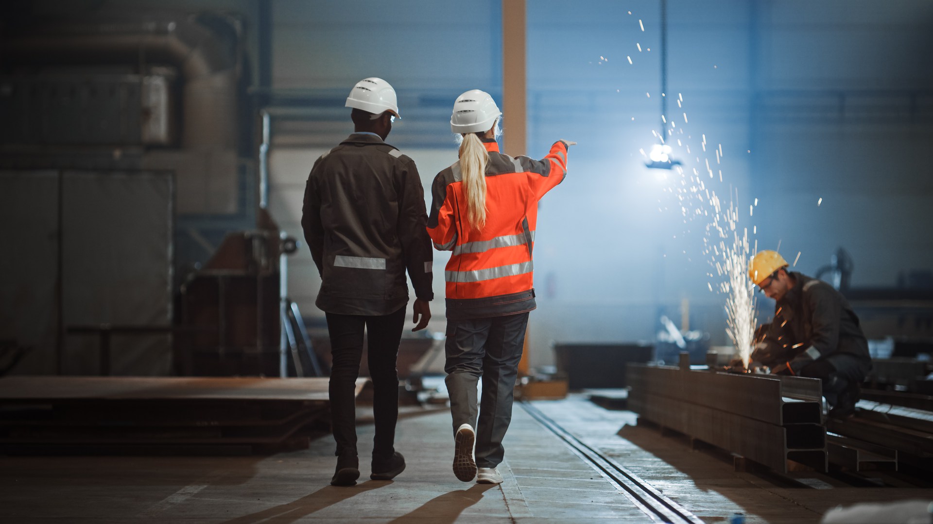 Two Heavy Industry Engineers Walk Away from Camera in Steel Factory and Discuss Work. Industrial Worker Uses Angle Grinder in the Background. African American Specialist Talks to Female Technician.